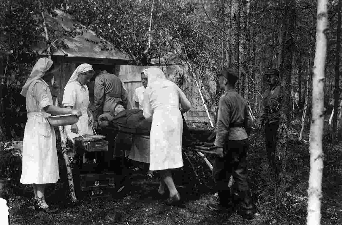 Neljä Punaisen Ristin sairaanhoitajaa työssään. Miestä tuodaan paareilla sisään kenttäsairaalaan.